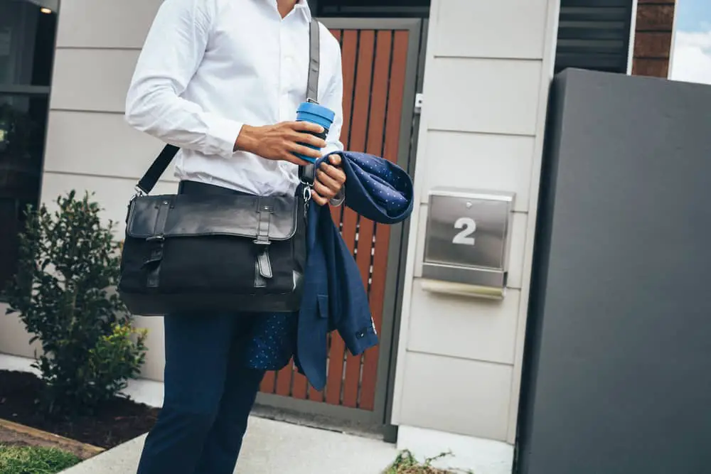 Young man standing outside his modern apartment with coffee and messenger bag